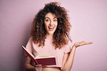 Young beautiful student woman with curly hair and piercing reading book over pink background very...