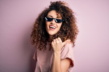 Young beautiful woman with curly hair and piercing wearing funny thug life sunglasses smiling with happy face looking and pointing to the side with thumb up.