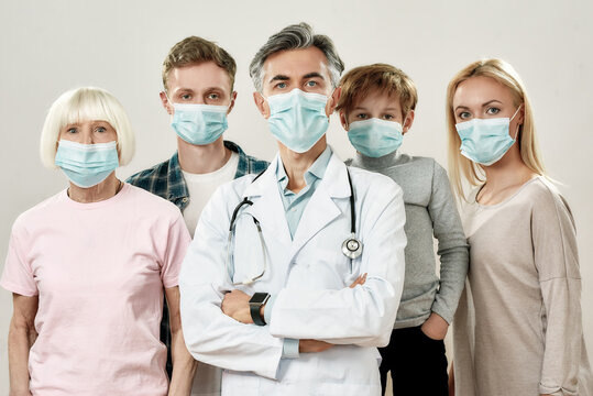 Big Family With Doctor. Portrait Of Mature Male Doctor And Family Of Four Wearing Medical Protective Masks, Looking At Camera While Standing Against Grey Background