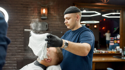 Traditional ritual of shaving. Side view of male barber with a mustache covering face of client...
