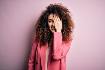 Young beautiful businesswoman with curly hair and piercing wearing elegant jacket covering one eye with hand, confident smile on face and surprise emotion.