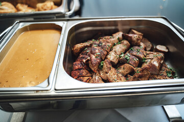 Stainless hotel pans on food warmers with various meals. Roasted pork meat pieces and creamy sauce. Self-service buffet table. Celebration, party