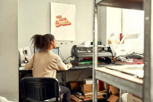 The total printing arrangement. Young woman sitting by computer monitor and sending chosen file to printing machine while printing advertisement sticker with text or picture for a T-shirt