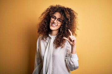 Young beautiful brunette woman with curly hair and piercing wearing shirt and glasses smiling and confident gesturing with hand doing small size sign with fingers looking and the camera. Measure