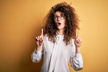 Young beautiful brunette woman with curly hair and piercing wearing shirt and glasses amazed and surprised looking up and pointing with fingers and raised arms.