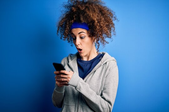 Beautiful sportswoman with curly hair and piercing wearing sportswear using smartphone scared in shock with a surprise face, afraid and excited with fear expression