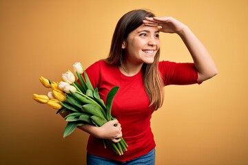 Young blonde woman holding romantic bouquet of tulips flowers over yellow background very happy and smiling looking far away with hand over head. Searching concept.