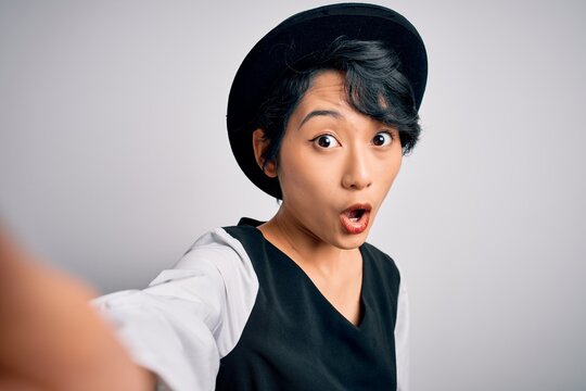 Beautiful asian girl wearing casual hat making selfie by the camera over white background scared in shock with a surprise face, afraid and excited with fear expression