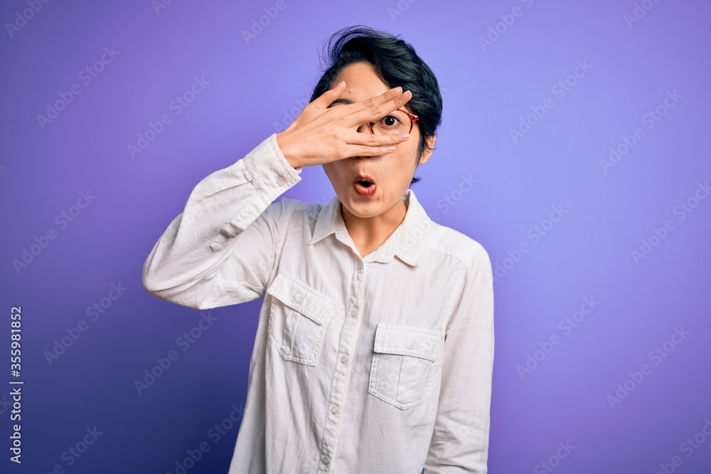 Poster Young beautiful asian girl wearing casual shirt and glasses standing over purple background peeking in shock covering face and eyes with hand, looking through fingers with embarrassed expression.