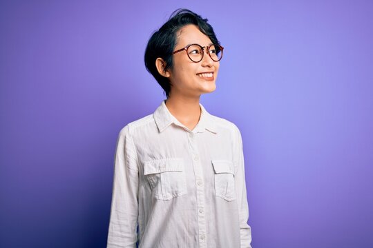 Young beautiful asian girl wearing casual shirt and glasses standing over purple background looking away to side with smile on face, natural expression. Laughing confident.