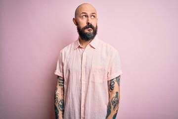 Handsome bald man with beard and tattoo wearing casual shirt over isolated pink background smiling looking to the side and staring away thinking.