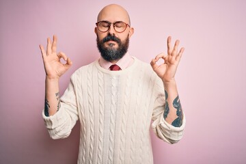 Handsome bald man with beard and tattoo wearing glasses and sweater over pink background relax and smiling with eyes closed doing meditation gesture with fingers. Yoga concept.