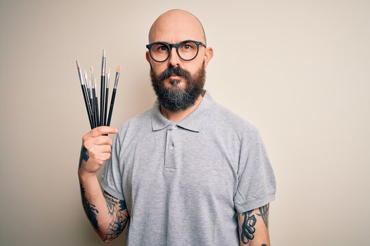 Handsome bald artist man with beard and tattoo painting using painter brushes with a confident expression on smart face thinking serious