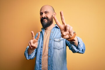 Handsome bald man with beard wearing casual denim jacket and striped t-shirt smiling looking to the camera showing fingers doing victory sign. Number two.