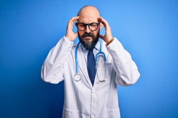 Handsome bald doctor man with beard wearing glasses and stethoscope over blue background with hand on headache because stress. Suffering migraine.