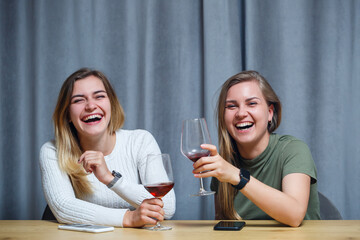 two girls of European appearance with blond hair are sitting at the table, drinking wine and laughing, relaxing at home, alcohol