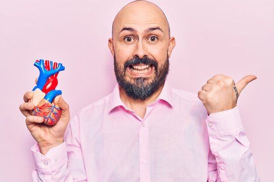 Young handsome man holding heart organ pointing thumb up to the side smiling happy with open mouth