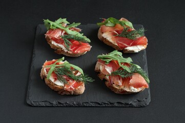 Buckwheat toasts with goat cheese, prosciutto and arugula lying on a black stone board on a black table. Closeup