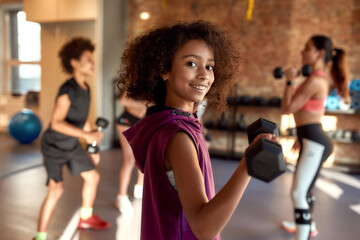 African american girl smiling at camera while exercising using dumbbell in gym together with female...