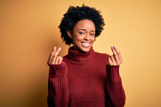 Young beautiful African American afro woman with curly hair wearing casual turtleneck sweater doing money gesture with hands, asking for salary payment, millionaire business