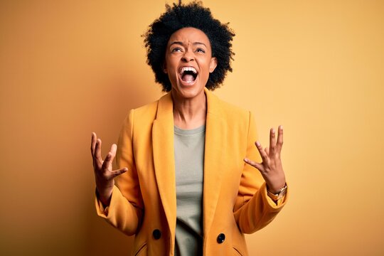 Young beautiful African American afro businesswoman with curly hair wearing yellow jacket crazy and mad shouting and yelling with aggressive expression and arms raised. Frustration concept.