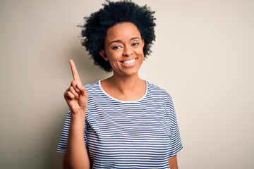 Young beautiful African American afro woman with curly hair wearing striped t-shirt showing and pointing up with finger number one while smiling confident and happy.