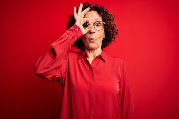 Middle age beautiful curly hair woman wearing casual shirt and glasses over red background doing ok gesture shocked with surprised face, eye looking through fingers. Unbelieving expression.