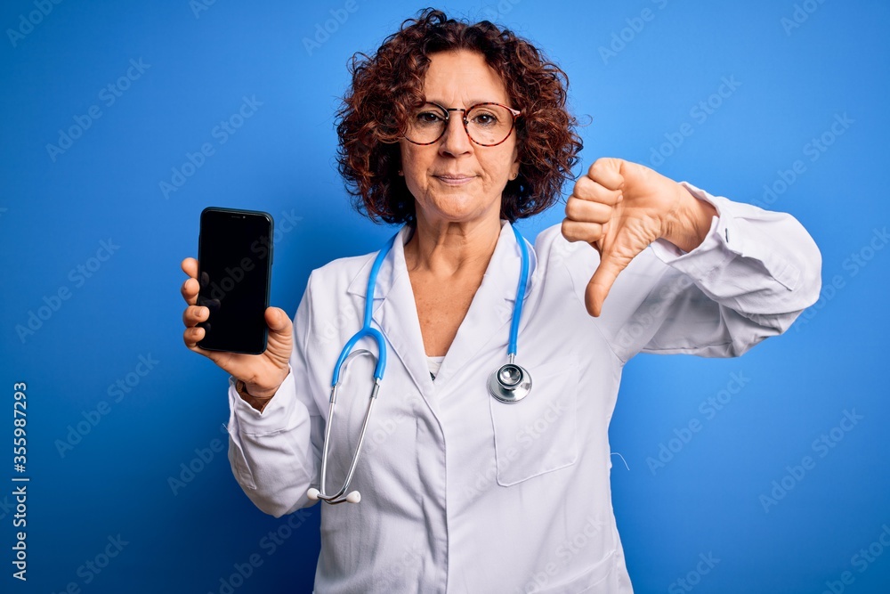 Sticker Middle age doctor woman wearing coat and stethoscope holding smartphone showing screen with angry face, negative sign showing dislike with thumbs down, rejection concept