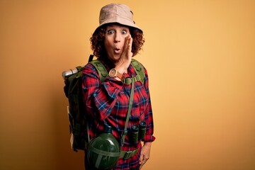 Middle age curly hair hiker woman hiking wearing backpack and water canteen using binoculars hand on mouth telling secret rumor, whispering malicious talk conversation
