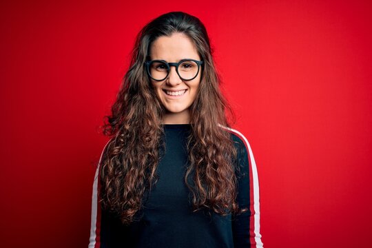 Young beautiful woman with curly hair wearing sweater and glasses over red background with a happy and cool smile on face. Lucky person.