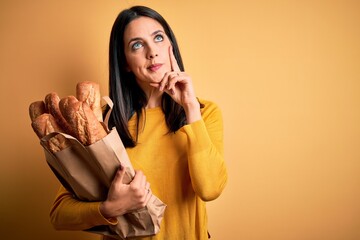 Young woman with blue eyes holding paper bag with bread over isolated yellow background serious face thinking about question, very confused idea