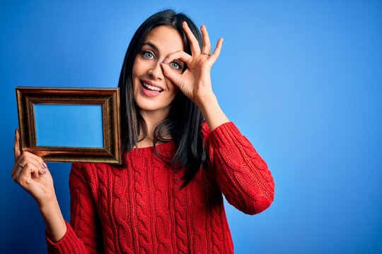 Young brunette woman with blue eyes holding empty photography frame over blue background with happy face smiling doing ok sign with hand on eye looking through fingers
