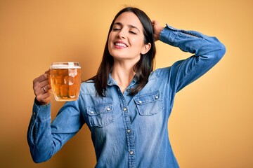 Young woman with blue eyes drinking jar of beer standing over isolated yellow background smiling confident touching hair with hand up gesture, posing attractive and fashionable