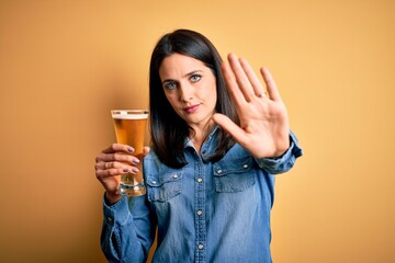 Young woman with blue eyes drinking glass of beer standing over isolated yellow background with open hand doing stop sign with serious and confident expression, defense gesture