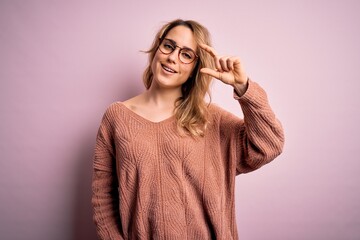 Young beautiful blonde woman wearing casual sweater and glasses over pink background smiling and confident gesturing with hand doing small size sign with fingers looking and the camera. Measure