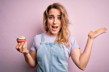 Young beautiful blonde woman eatimg chocolate cupcake over isolated pink background very happy and excited, winner expression celebrating victory screaming with big smile and raised hands