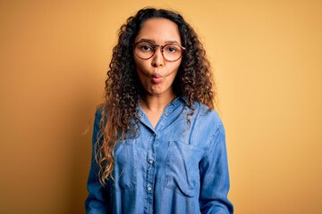 Beautiful woman with curly hair wearing casual denim shirt and glasses over yellow background making fish face with lips, crazy and comical gesture. Funny expression.