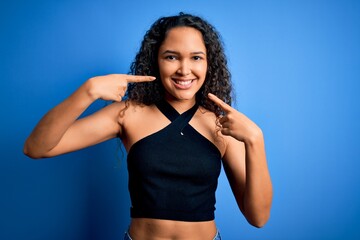 Young beautiful woman with curly hair wearing casual t-shirt standing over blue background smiling cheerful showing and pointing with fingers teeth and mouth. Dental health concept.