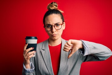 Young beautiful blonde businesswoman with blue eyes wearing glasses drinking cup of coffe with angry face, negative sign showing dislike with thumbs down, rejection concept