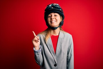 Young beautiful blonde motorcyclist woman wearing motorcycle helmet over red background with a big smile on face, pointing with hand and finger to the side looking at the camera.