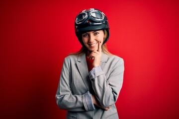 Young beautiful blonde motorcyclist woman wearing motorcycle helmet over red background looking confident at the camera with smile with crossed arms and hand raised on chin. Thinking positive.