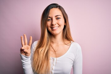 Young beautiful blonde woman with blue eyes wearing white t-shirt over pink background showing and pointing up with fingers number three while smiling confident and happy.