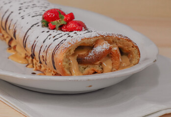 Sponge cake with tea on a wooden table.
One biscuit roll in a white plate lies on a wooden table with a white napkin from below, side view close-up.