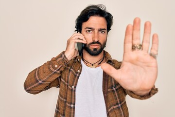 Young handsome hispanic man having a conversation talking on smartphone with open hand doing stop sign with serious and confident expression, defense gesture