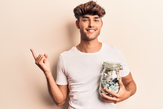 Young hispanic man holding jar with savings smiling happy pointing with hand and finger to the side