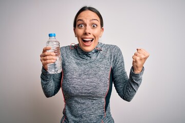 Young fitness woman wearing sport workout clothes drinking water from plastic bottle screaming proud and celebrating victory and success very excited, cheering emotion