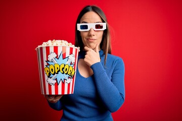 Young blonde woman wearing 3d glasses and eating pack of popcorn watching a movie on cinema looking confident at the camera smiling with crossed arms and hand raised on chin. Thinking positive.