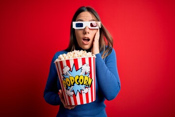 Young blonde woman wearing 3d glasses and eating pack of popcorn watching a movie on cinema afraid and shocked, surprise and amazed expression with hands on face