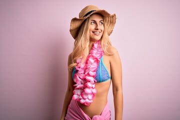 Young beautiful blonde woman on vacation wearing bikini and hat with hawaiian lei flowers looking away to side with smile on face, natural expression. Laughing confident.