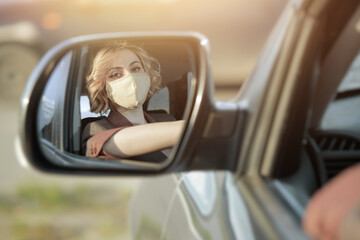 Beautiful girl driver with a mask on her face, sitting at the wheel of the car reflected in the side mirror of the car at sunset.